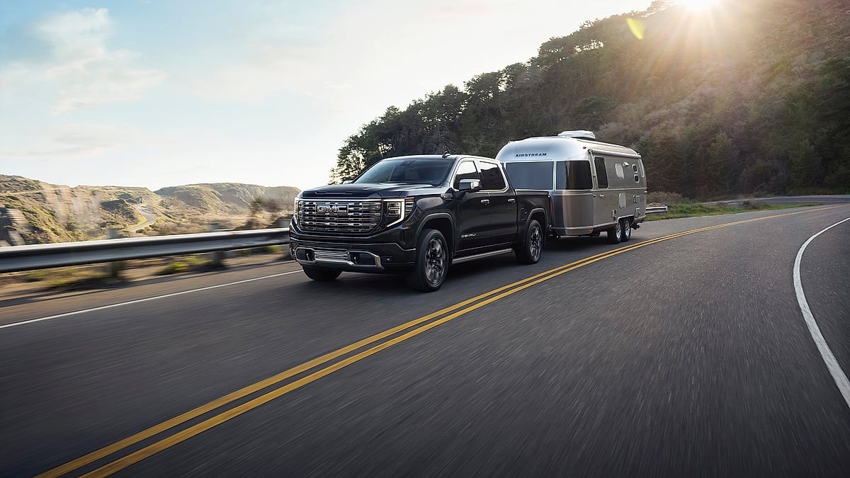 Black GMC Sierra Denali Ultimate towing an Airstream camper on a winding coastal road at sunset.