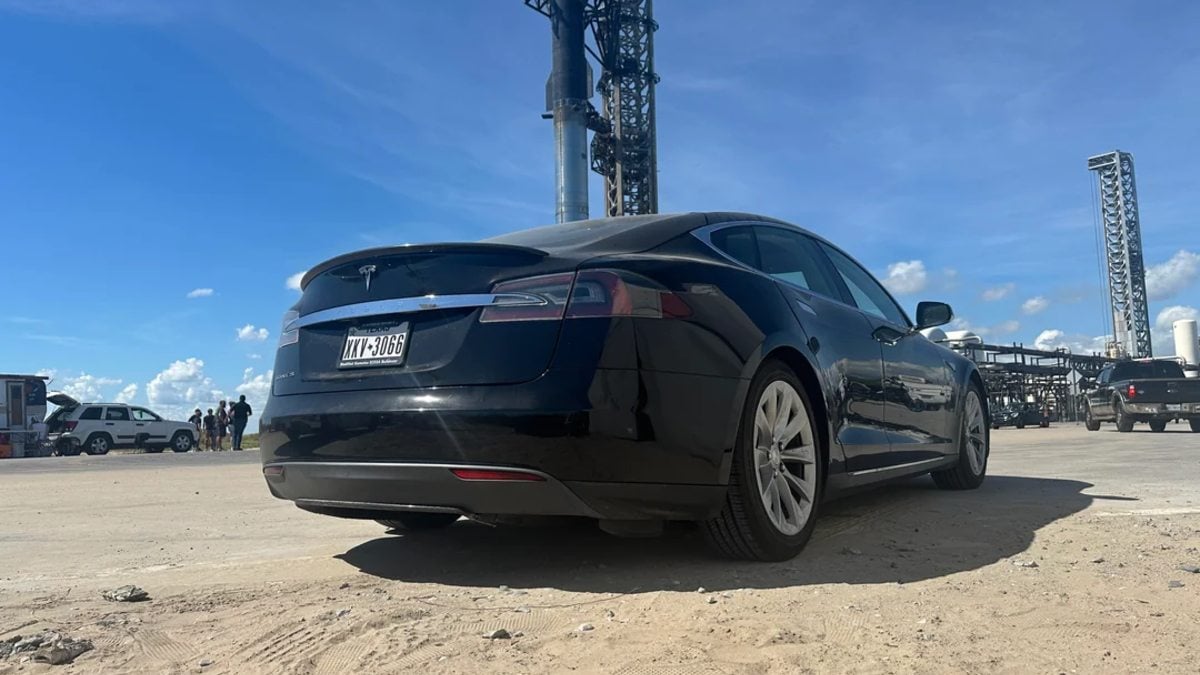 Black Tesla car parked on a dirt lot near industrial structures under a clear blue sky.