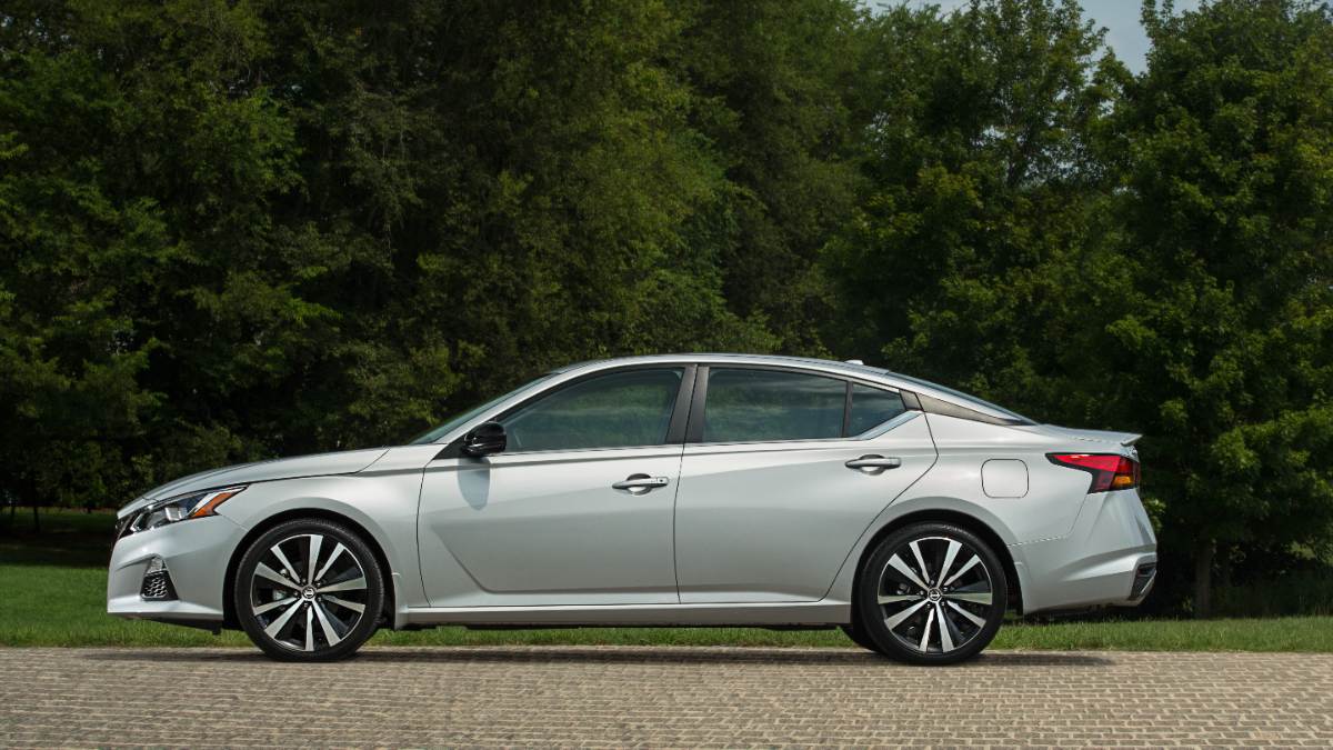 Silver 2019 Nissan Altima sedan parked on brick pathway with lush green trees in background, side profile view