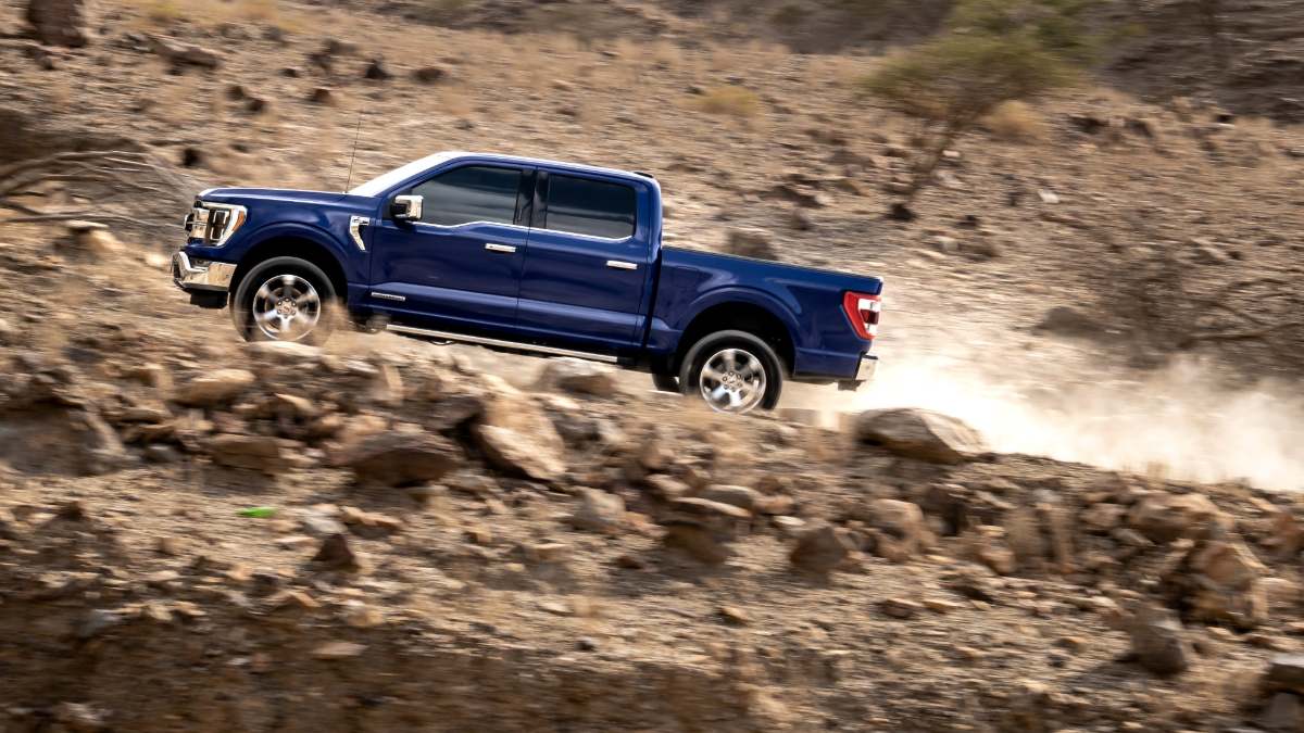 A blue 2021 Ford F-150 pickup truck shown in profile view climbing a steep rocky incline, demonstrating its off-road capabilities in a dynamic action shot.
