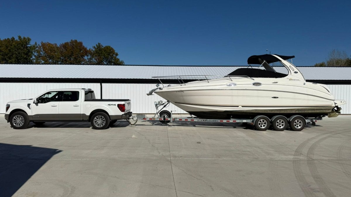 White Ford F-150 pickup truck towing a large Sea Ray cabin cruiser boat on triple-axle trailer, parked outside storage facility with metal siding.