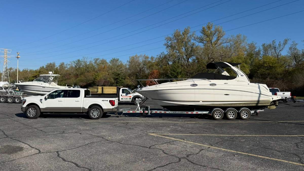 White Ford F-150 4x4 pickup truck towing a large Sea Ray cabin cruiser boat on triple-axle trailer, parked in marina lot under blue sky.