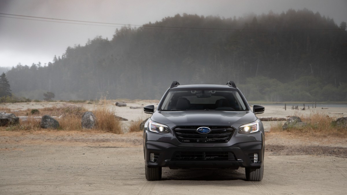 A dark gray 2021 Subaru Outback is shown from the front angle, parked on a misty beach or shoreline with forested hills in the background.
