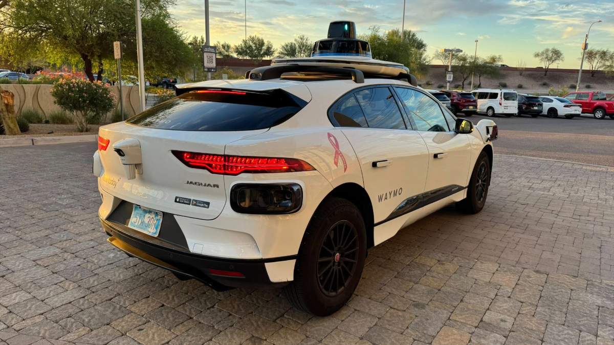 White Waymo autonomous Jaguar i-Pace with rooftop sensors, parked on brick pavement at sunset with trees and parking lot in background