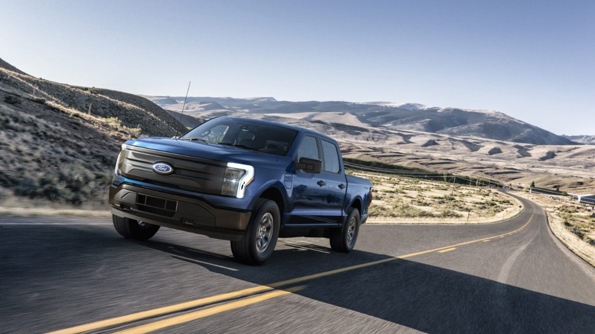 2022 Ford F-150 Lightning electric pickup truck in dark blue, shown at three-quarter front angle climbing mountainous road, featuring distinctive LED light bar and black grille.