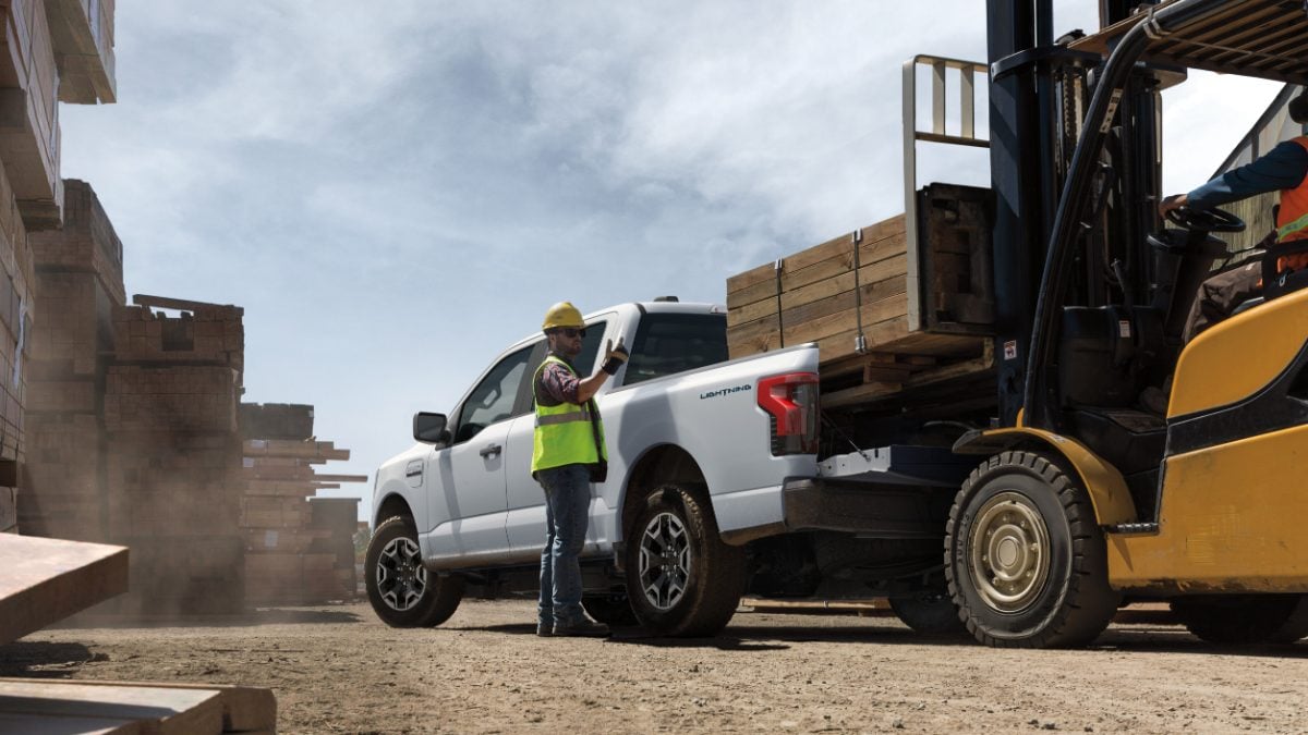 A white Ford F-150 Lightning EV pickup truck is shown from the side at a construction or warehouse site, with a yellow forklift visible in the foreground.