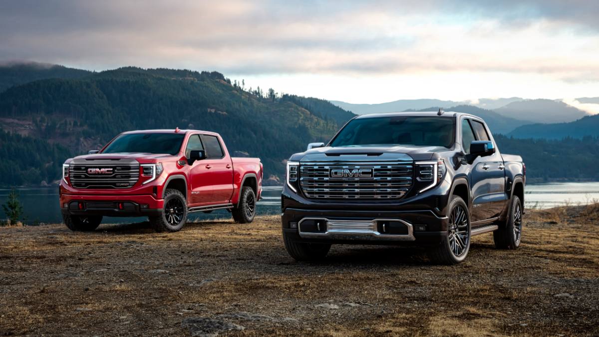 Two GMC trucks parked on a dirt area with mountains and a lake in the background.