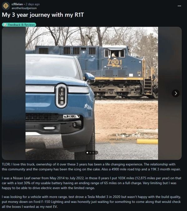 A close-up of a blue Rivian R1T electric truck with a prominent front design, parked by a railroad track. A train is visible in the background.