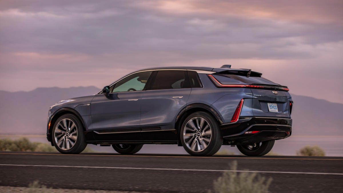 Rear three-quarter view of a 2023 Cadillac Lyriq electric SUV driving at sunset, highlighting sleek taillights and modern crossover design.