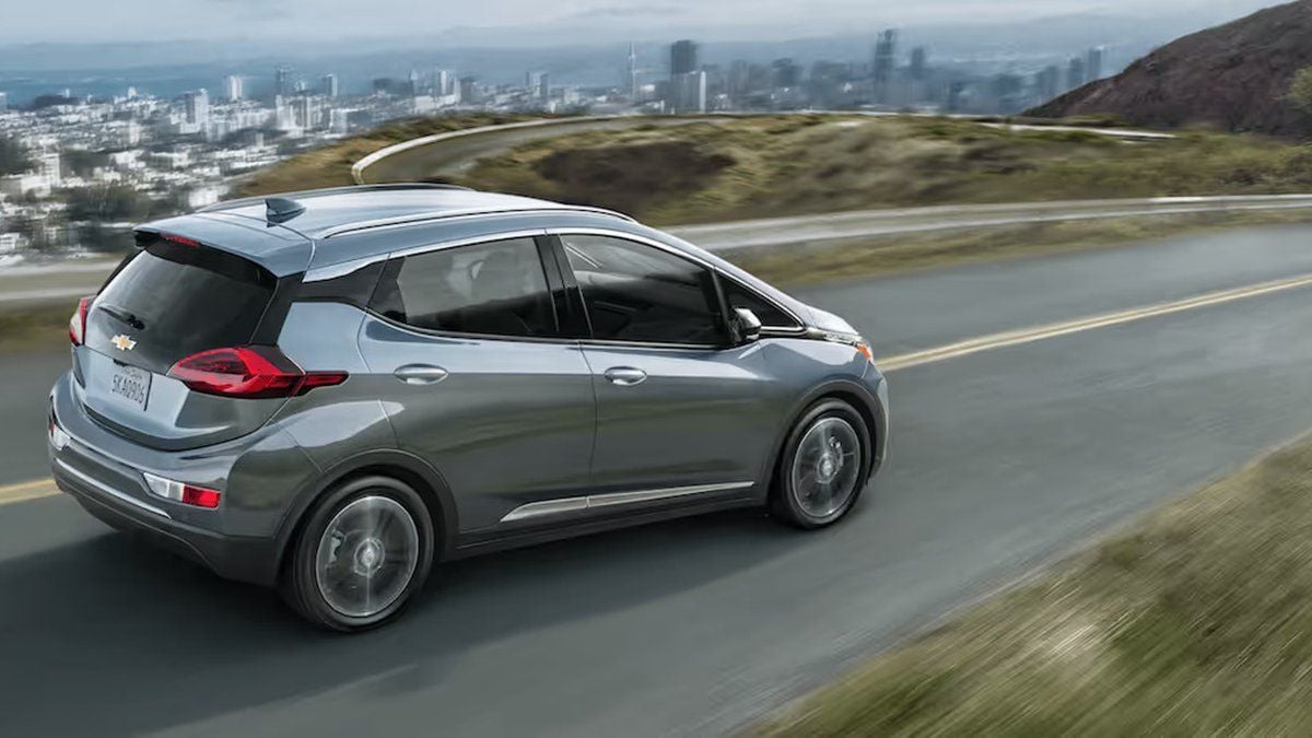 A silver Chevy Bolt EV drives along a winding road. The city skyline is visible in the background, conveying a sense of freedom and adventure.