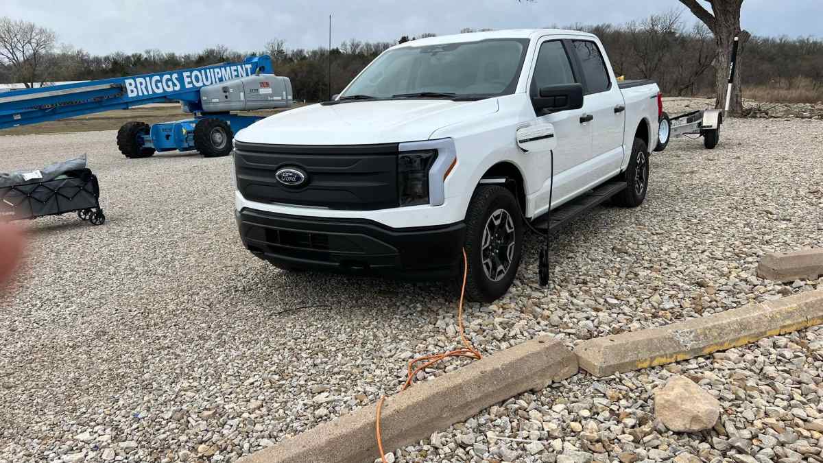 A white 2023 Ford F-150 Lightning electric pickup truck shown from a front three-quarter angle, parked on gravel with construction equipment visible in the background.