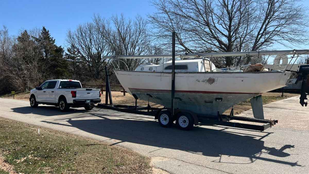 A white sailboat on a dual-axle trailer being towed by a white pickup truck, photographed from the side on a sunny day with bare winter trees in the background.