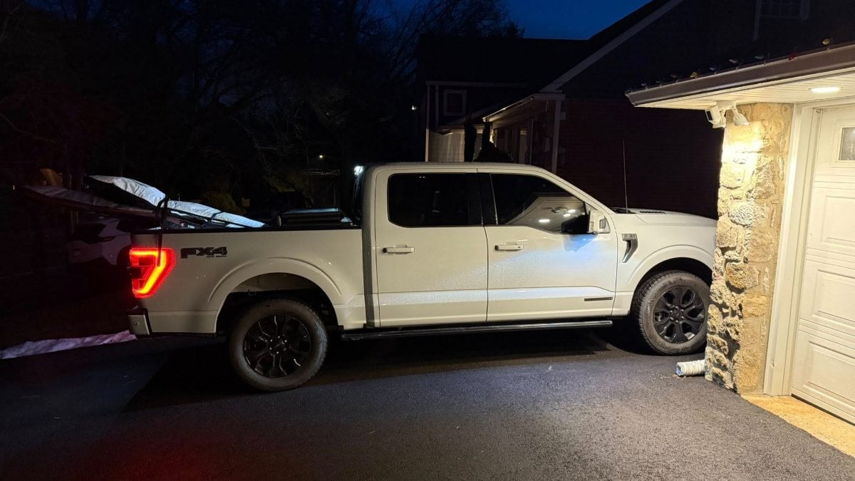 White Ford F-150 pickup truck parked in driveway at night, illuminated by garage lights, featuring black wheels and distinctive LED taillights.