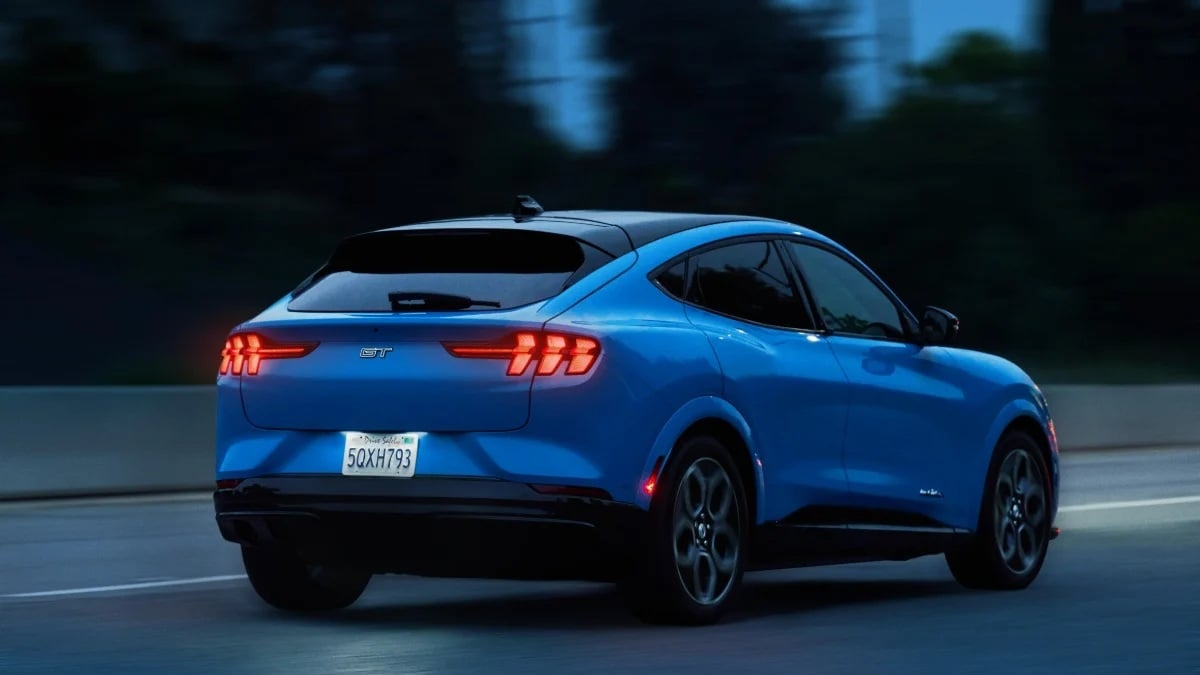 Blue 2023 Ford Mustang Mach-E GT driving on the highway at dusk, rear three-quarter view showing LED taillights and sporty electric SUV design.