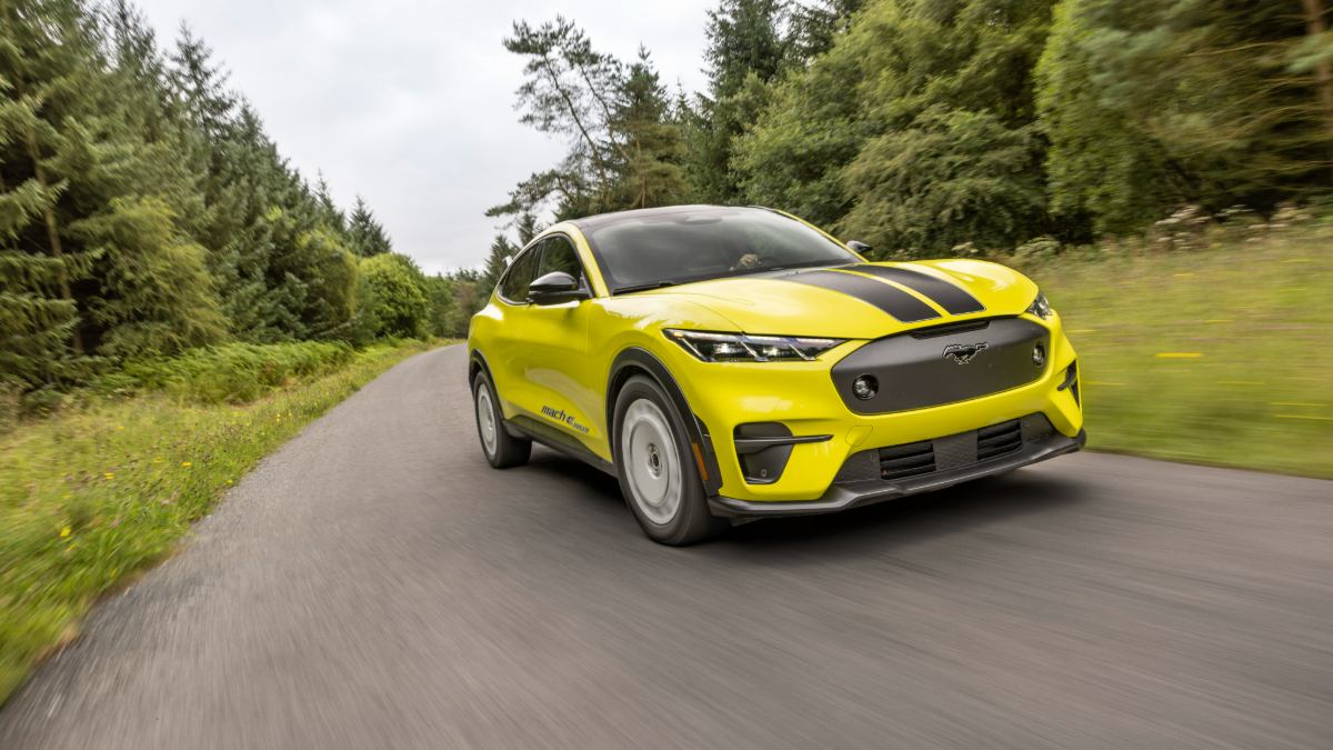 A bright yellow 2023 Ford Mustang Mach-E rally car shown from the front three-quarter view, captured mid-jump on a dirt road with a forested background.