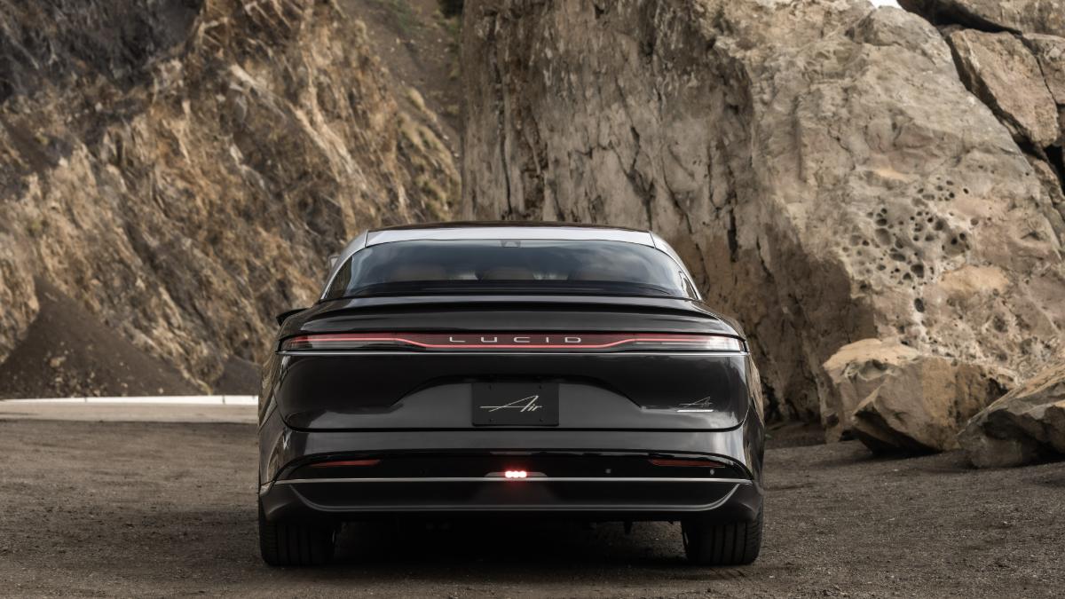 A black Lucid Air luxury electric sedan shown from the rear view, featuring its distinctive full-width LED light bar and sleek fastback design, photographed against a dramatic rocky cliff backdrop.