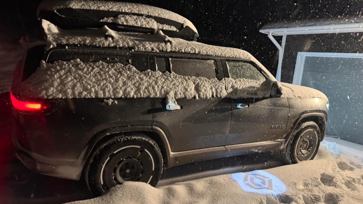 A black Rivian R1S SUV covered in fresh snow, photographed from the side at night with its tail lights illuminated.