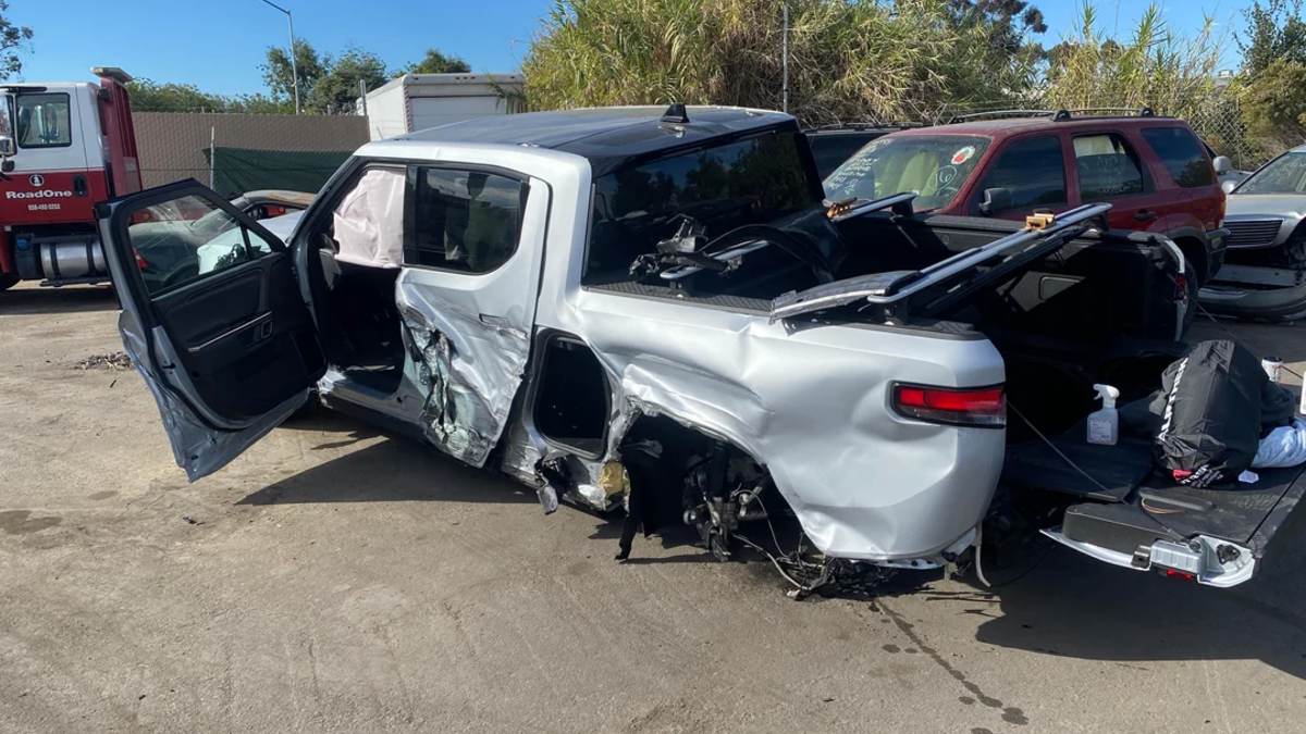 A severely damaged silver Rivian R1T shown from the rear quarter view, with significant impact damage to the passenger side, appears to be in a salvage or tow yard.