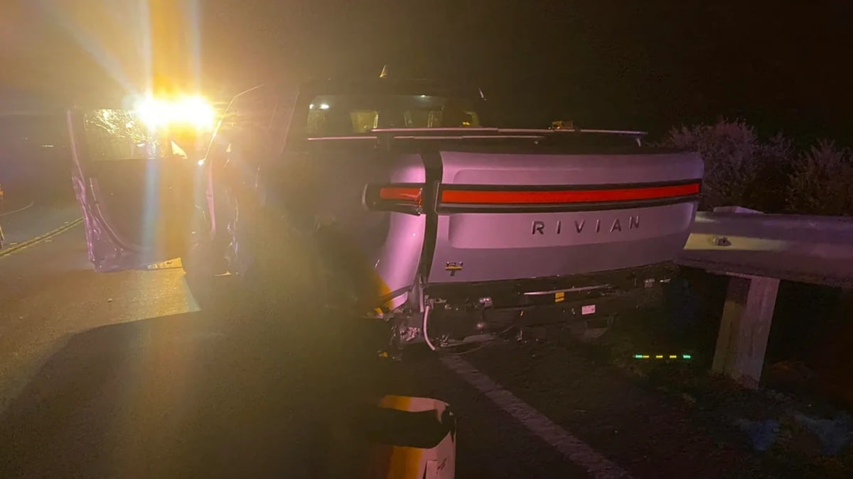 A nighttime view of a Rivian electric vehicle's damaged rear end and badging, illuminated in red with ambient lighting creating a moody atmosphere.