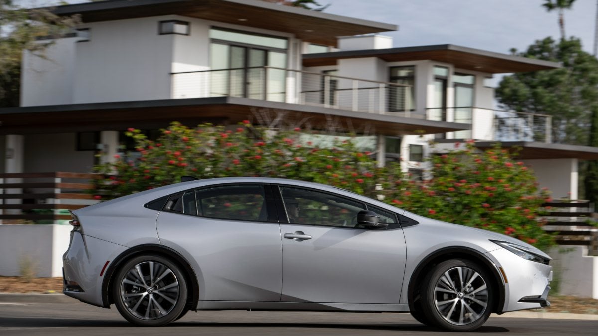 A side view of a silver 2023 Toyota Prius driving past modern houses.
