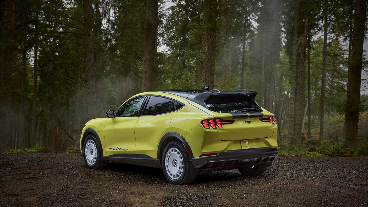 Rear three-quarter view of the 2023 Ford Mustang Mach-E Rally in a wooded setting, showing raised suspension, rear spoiler, and off-road-inspired design.