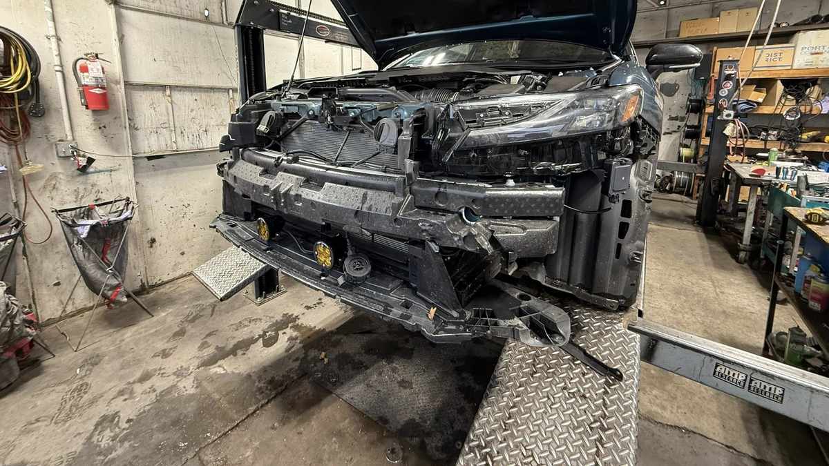 Close-up front view of a 2023 Toyota Camry in Guard Metallic on a hydraulic lift in a repair shop, showing the front bumper, grille, and auxiliary yellow fog lights.