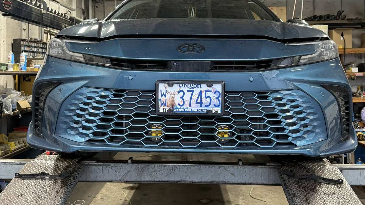 2023 Toyota Camry in Guard Metallic on a lift in a repair shop with the hood open and front bumper removed, exposing the internal front end components.