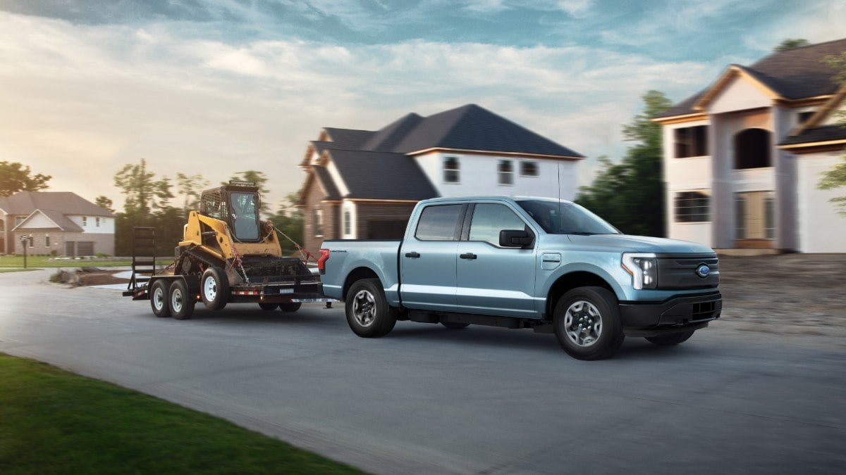 Light blue 2022 Ford F-150 Lightning electric pickup truck shown from the front 3/4 angle, towing construction equipment on a trailer in a residential neighborhood setting.