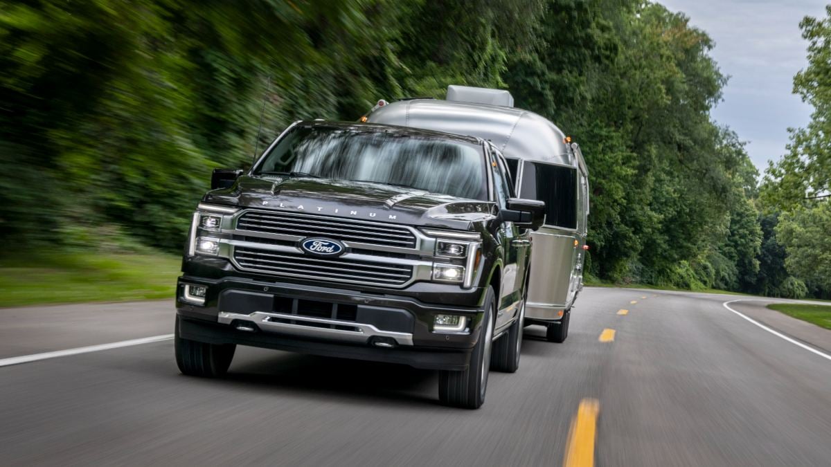 A dark gray 2024 Ford F-150 Platinum is shown from the front 3/4 angle towing an Airstream travel trailer on a curved road surrounded by green trees.