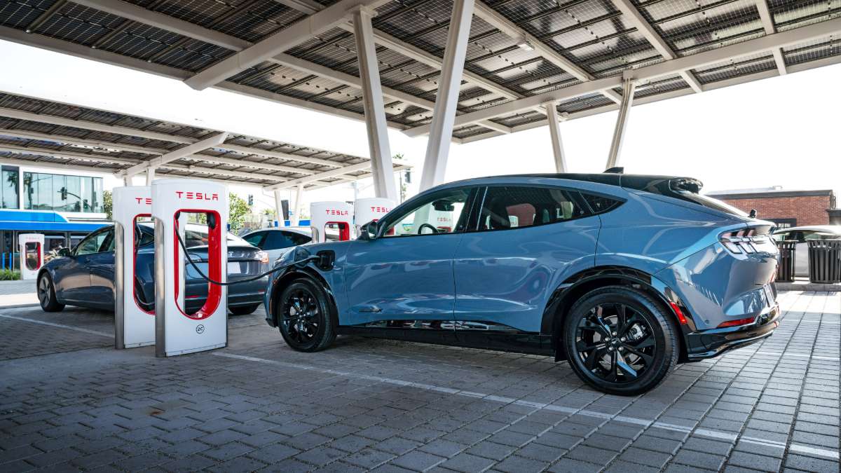 A blue Ford Mustang Mach-E is parked at a Tesla Supercharger station, shown from the rear three-quarter view with solar panels visible overhead on the charging station canopy.
