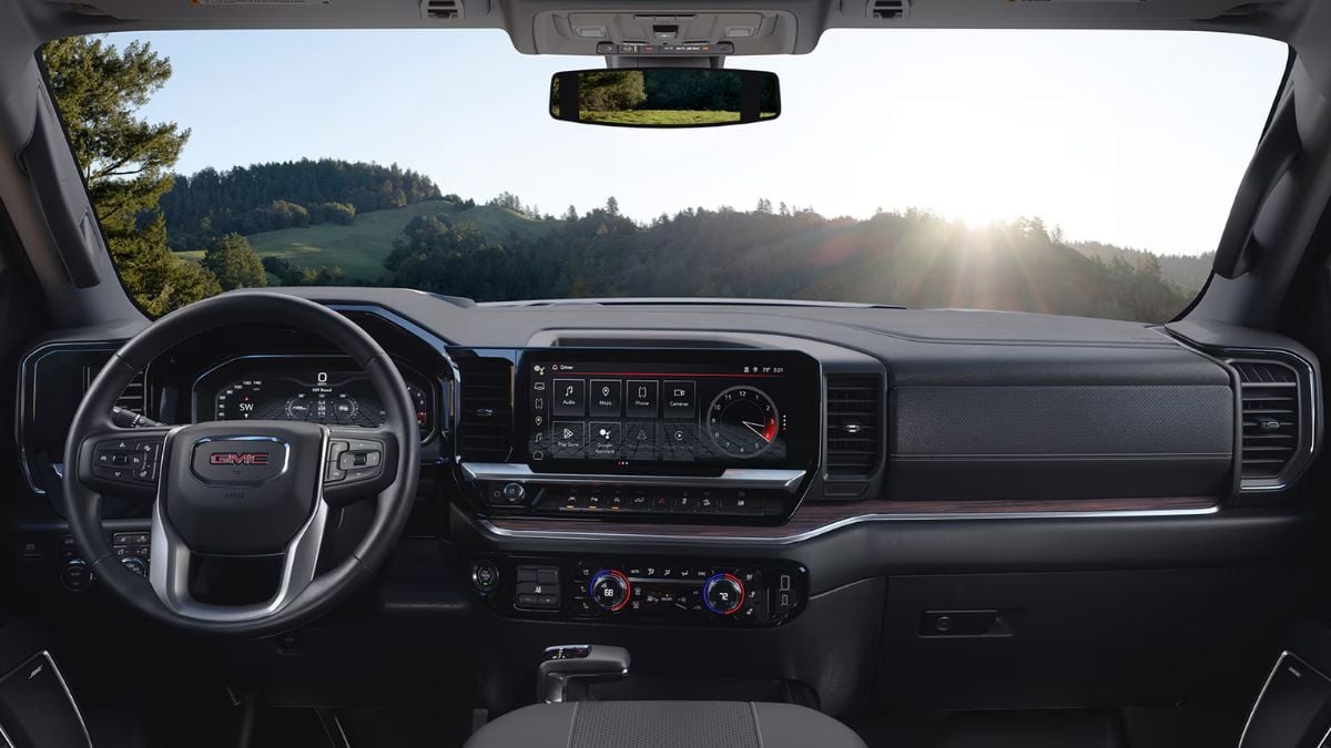 Inside view of a GMC Sierra 1500’s modern dashboard with a steering wheel, digital display screens, and various controls. Sunlit hills are visible through the windshield