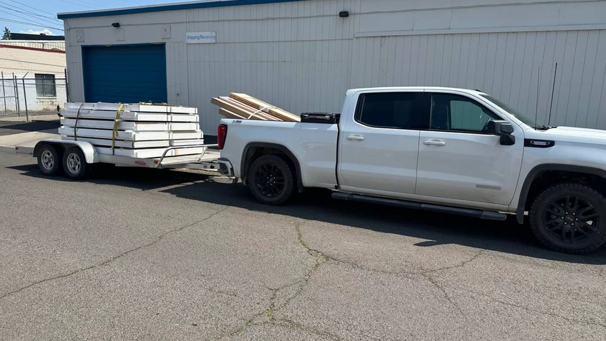 White 2024 GMC Sierra pickup truck with black wheels parked outside warehouse, towing flatbed trailer loaded with construction materials.