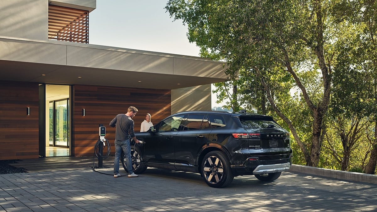 A couple charges a 2024 Honda Prologue in a modern driveway surrounded by trees. The house's sleek wooden facade complements the eco-friendly scene.