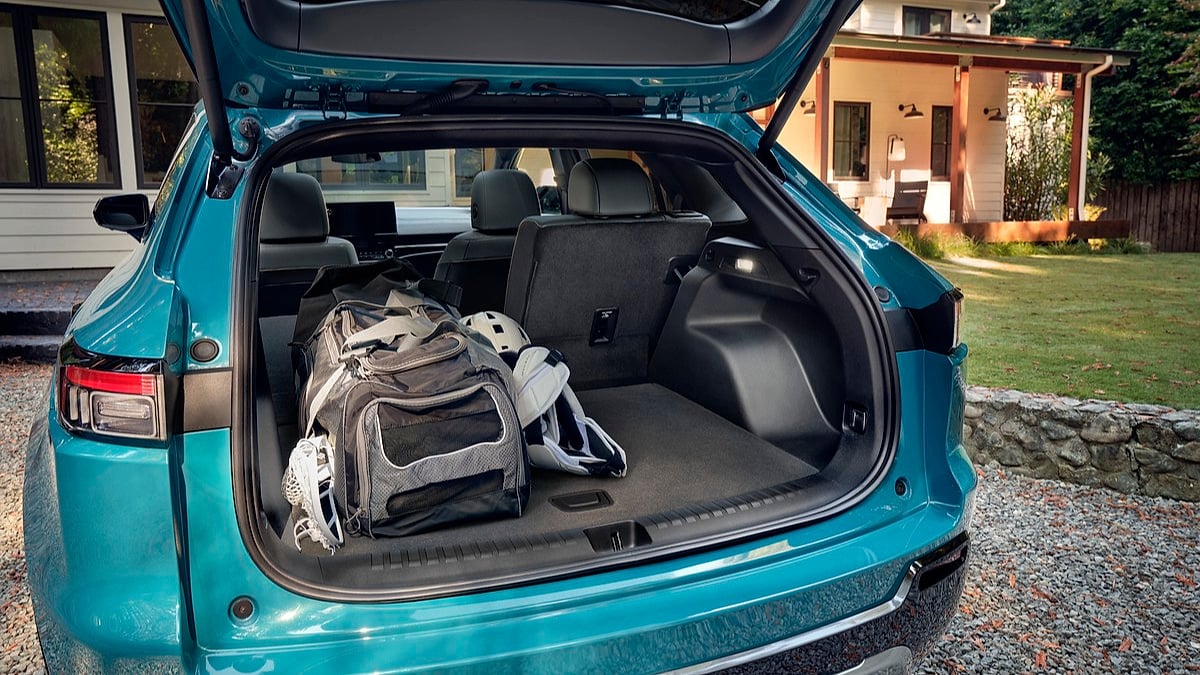 Open trunk of a blue 2024 Honda Prologue contains a sports bag and helmet, parked on a gravel driveway in front of a modern house surrounded by greenery.