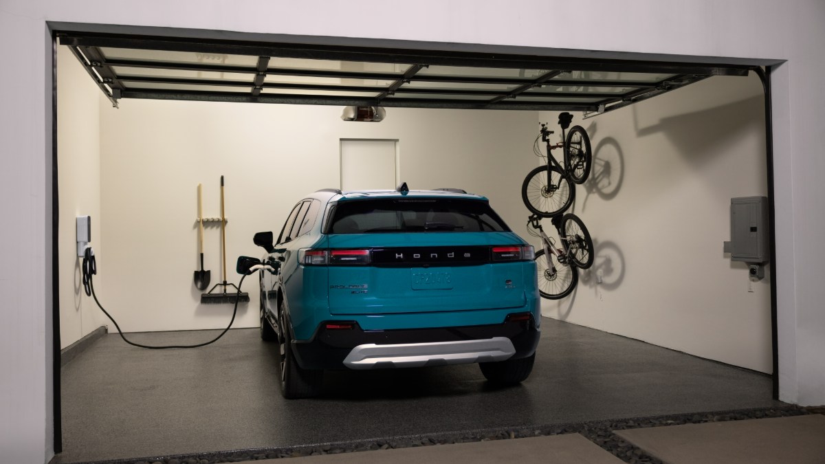 2024 Honda Prologue in turquoise blue, rear three-quarter view, parked in home garage with EV charger and wall-mounted bicycle.