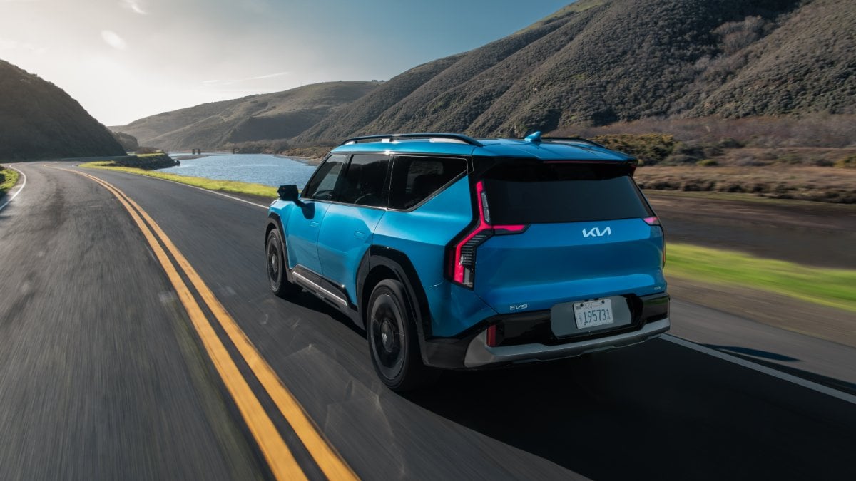A bright blue 2024 Kia EV9 electric SUV shown from the rear three-quarter angle, driving on a scenic coastal mountain road with water and hills visible in the background.