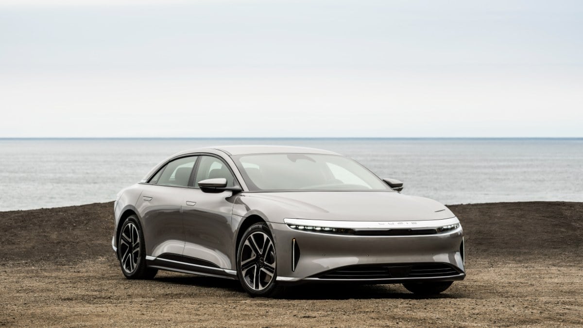 A silver Lucid Air luxury electric sedan shown from a front three-quarter angle, parked on a coastal terrain with the ocean visible in the background.