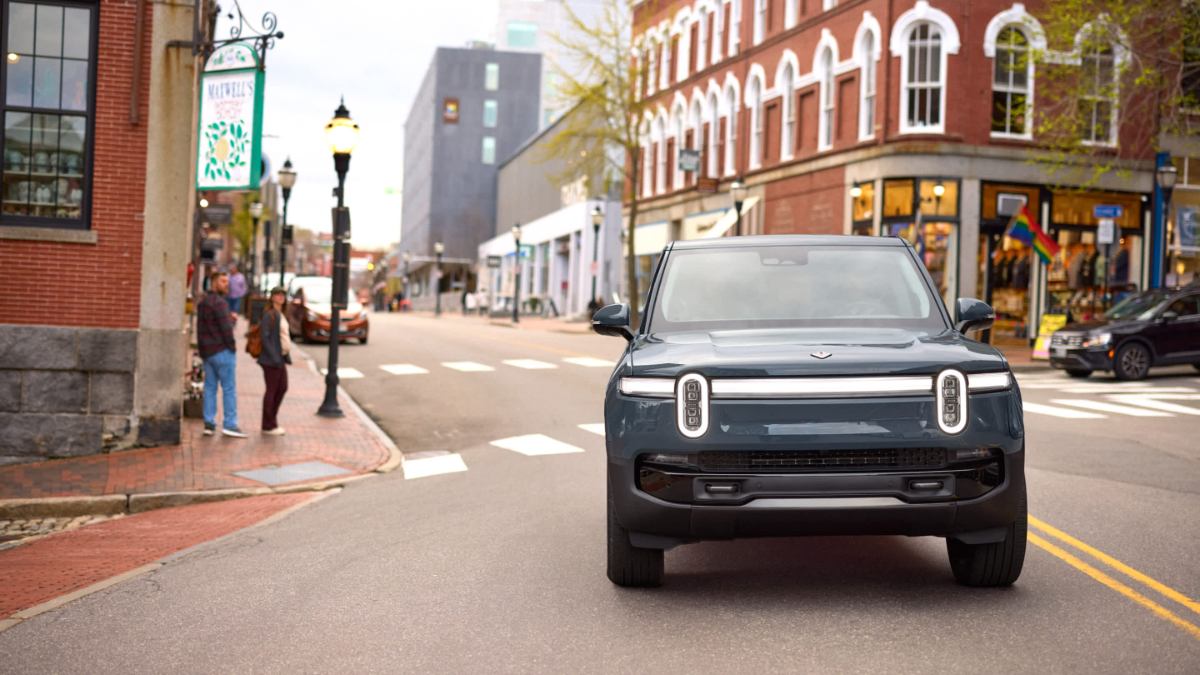 Dark blue Rivian R1S electric SUV photographed from front view on urban street, featuring distinctive vertical LED headlights and brick buildings.