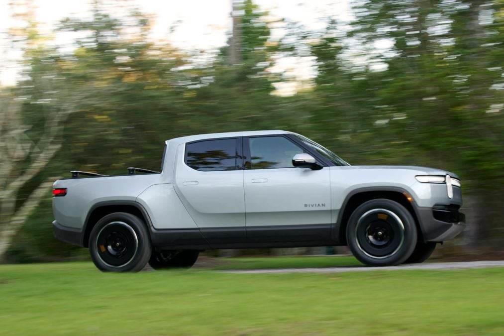 Side profile of a gray Rivian R1T electric pickup truck driving through a green wooded area, showcasing the all-electric adventure truck’s modern design, black wheels, and crew cab configuration.