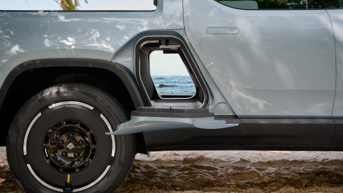 Close-up side view of a silver Rivian R1T's wheel and fender area, featuring the distinctive black wheel design with metallic accents against an oceanfront background.