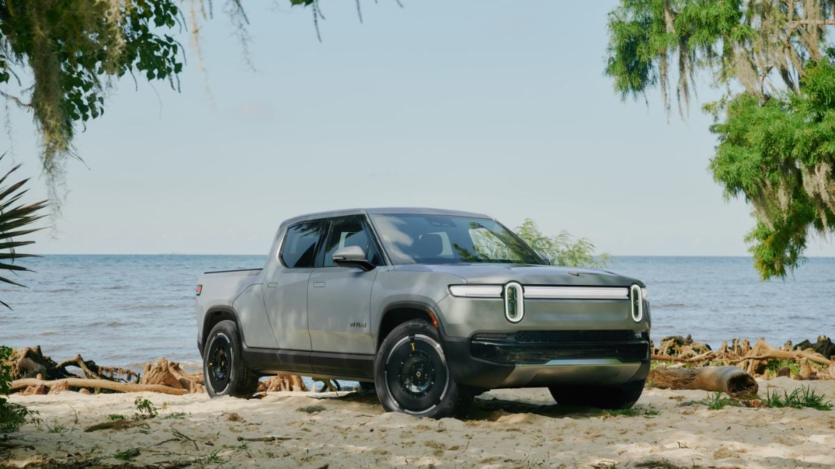 Silver Rivian R1T electric pickup truck parked on sandy beach with ocean view, framed by hanging moss and trees, showcasing distinctive front light bar design.