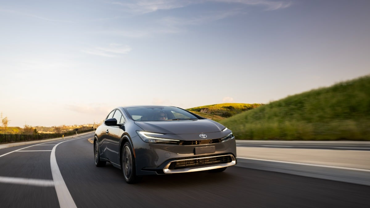 2023 Toyota Prius shown in dark gray, front three-quarter view, driving on curved mountain road at sunset, featuring sleek aerodynamic design and LED lighting.
