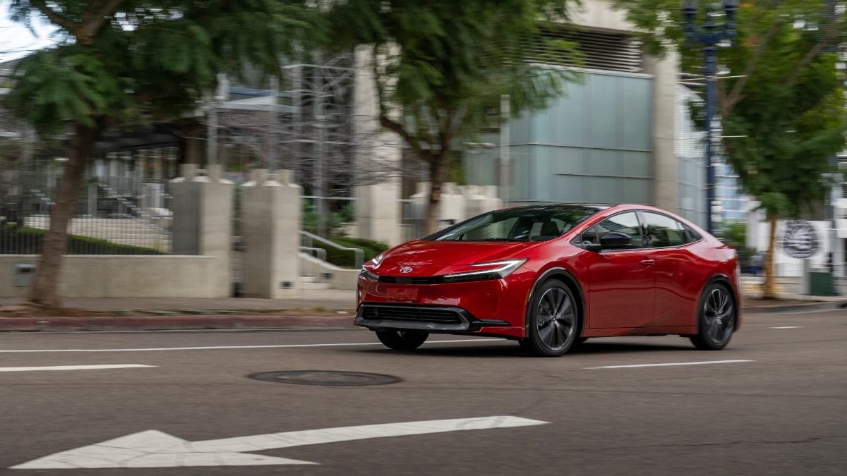 A red 2024 Toyota Prius shown from a rear three-quarter angle, driving on an urban street with trees in the background, showcasing its sleek new redesigned body style.