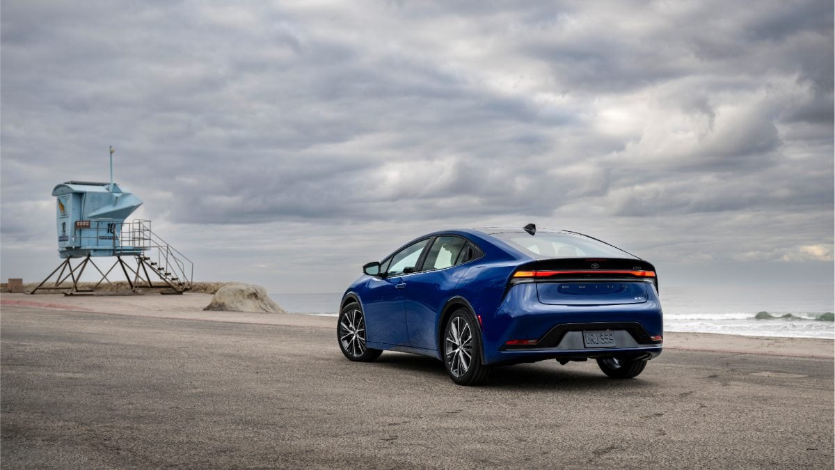 A blue 2024 Toyota Prius shown from the rear three-quarter view, parked on a beach with a lifeguard tower and dramatic cloudy sky in the background.