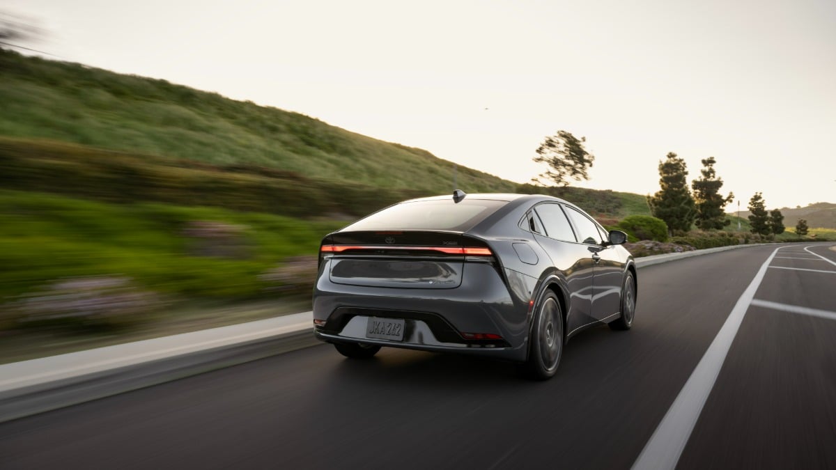 2023 Toyota Prius in metallic gray, rear three-quarter view, driving on winding mountain road at sunset, featuring distinctive fastback design and full-width LED taillights.