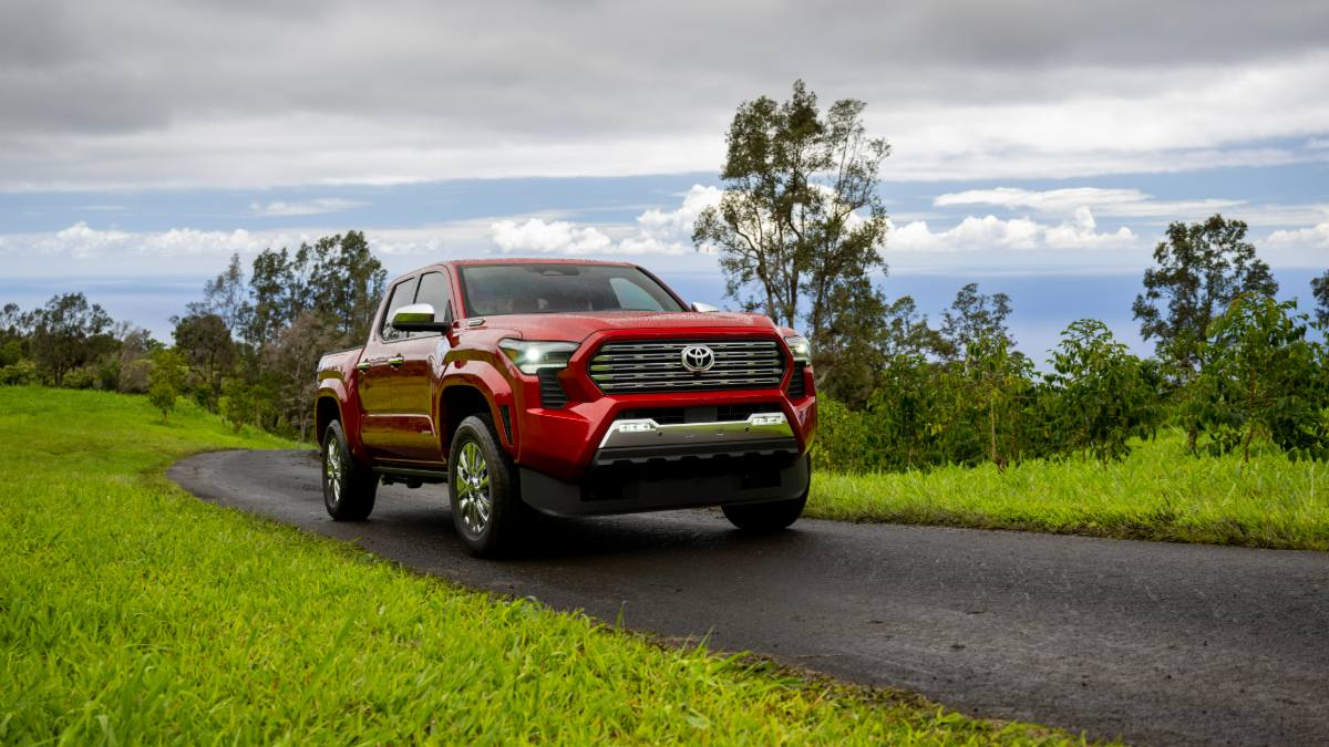 A red 2024 Toyota Tundra pickup truck shown from the front three-quarter view, parked on a winding rural road surrounded by lush green grass and trees under a partly cloudy sky.