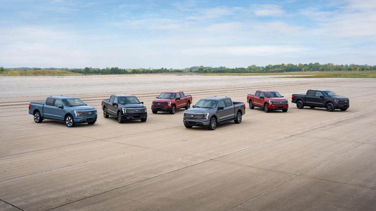 A group of six Ford F-150 Lightning electric pickup trucks in various colors (blue, gray, red, black) parked in a line on a large airport tarmac under a blue sky.