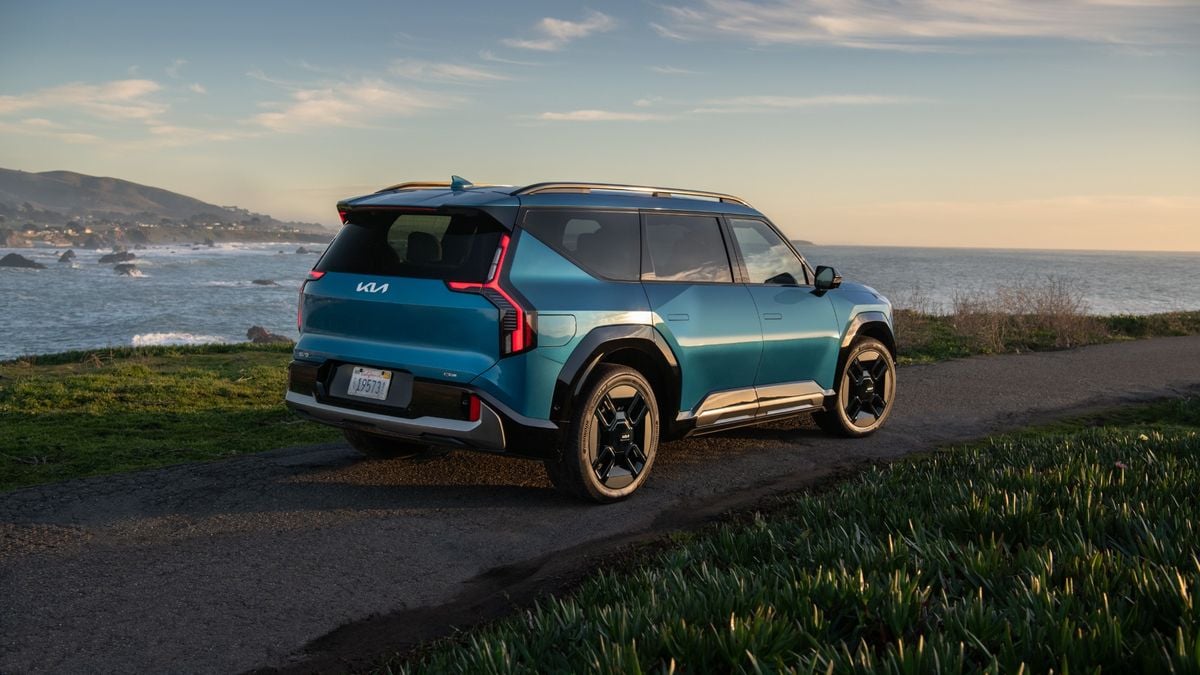 Blue 2024 Kia EV9 electric SUV parked on a coastal road at sunset with ocean and cliffs in the background.