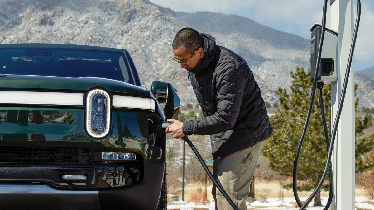 Blue Rivian R1S electric SUV charging at a public EV charging station with a person plugging in the charging cable.