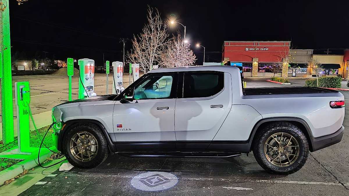 White 2024 Rivian R1T electric pickup charging at a public EV station at night.